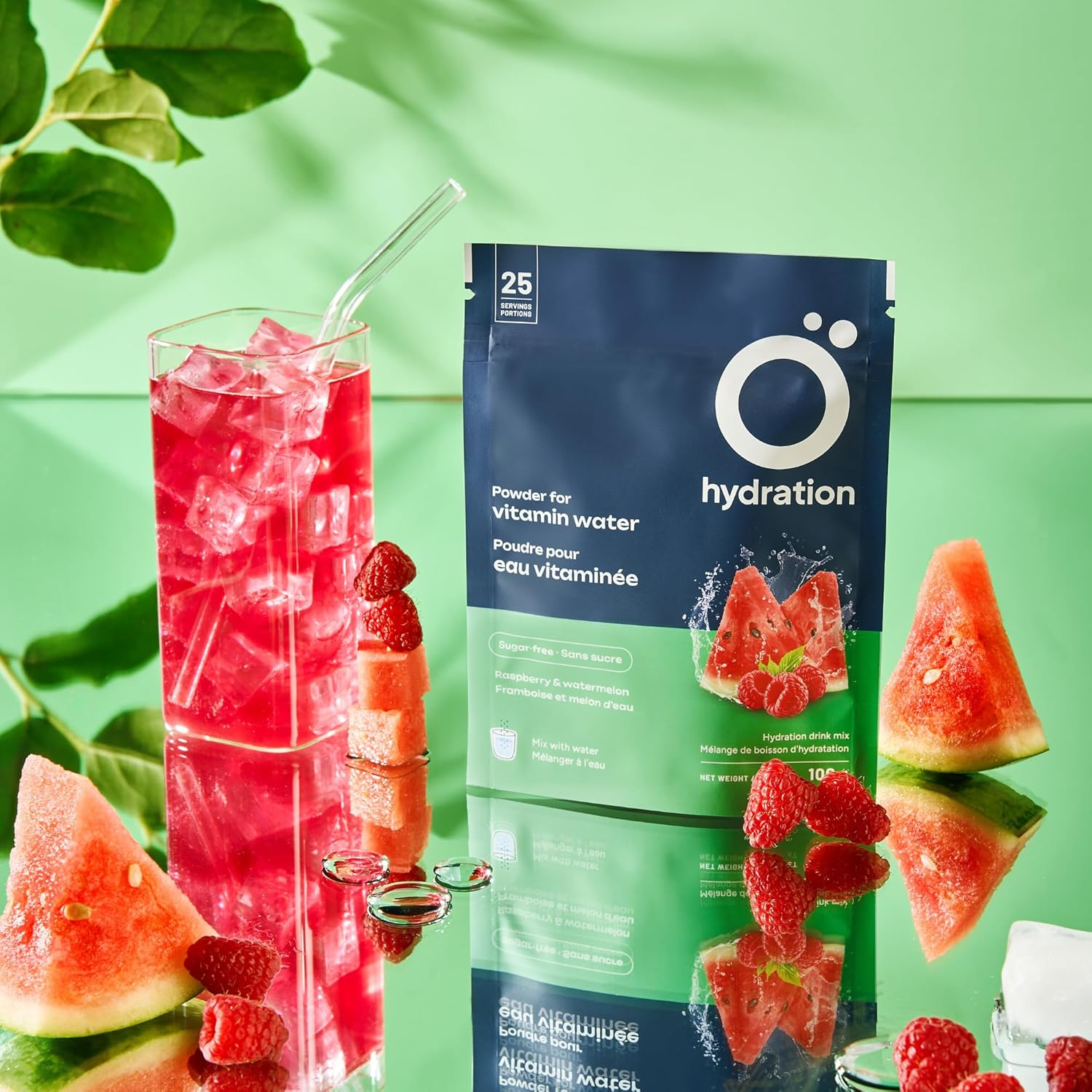 Ö Hydration Vitamized Powder 100g in raspberry & watermelon flavor, beside a glass of pink vitamin water, surrounded by fresh fruit and reflected on a glossy surface.